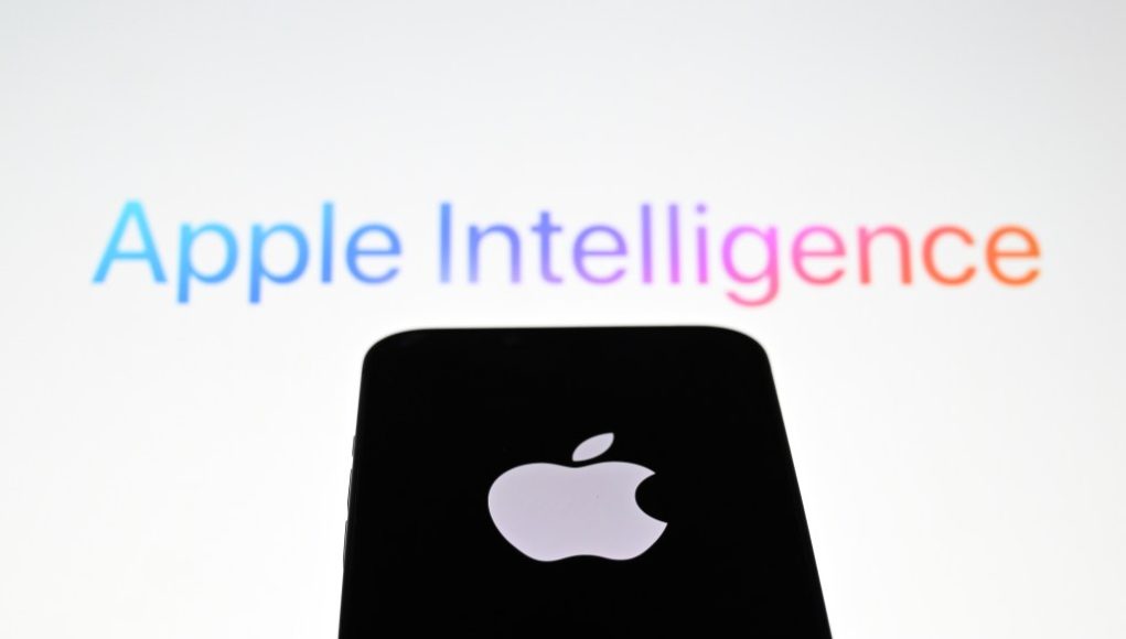 In this photo illustration, the 'Apple' logo is displayed on a mobile phone screen in front of a computer screen displaying Apple Intelligence logo.